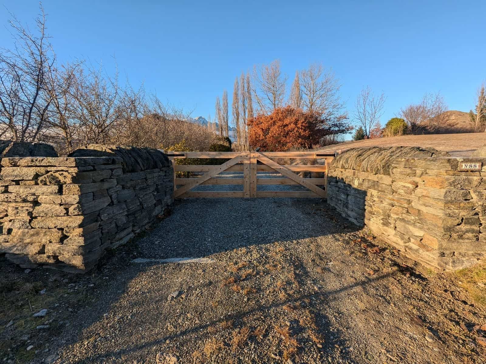 Macrocarpa 5-bar gate flanked by schist stone walls under blue sky, Central Otago